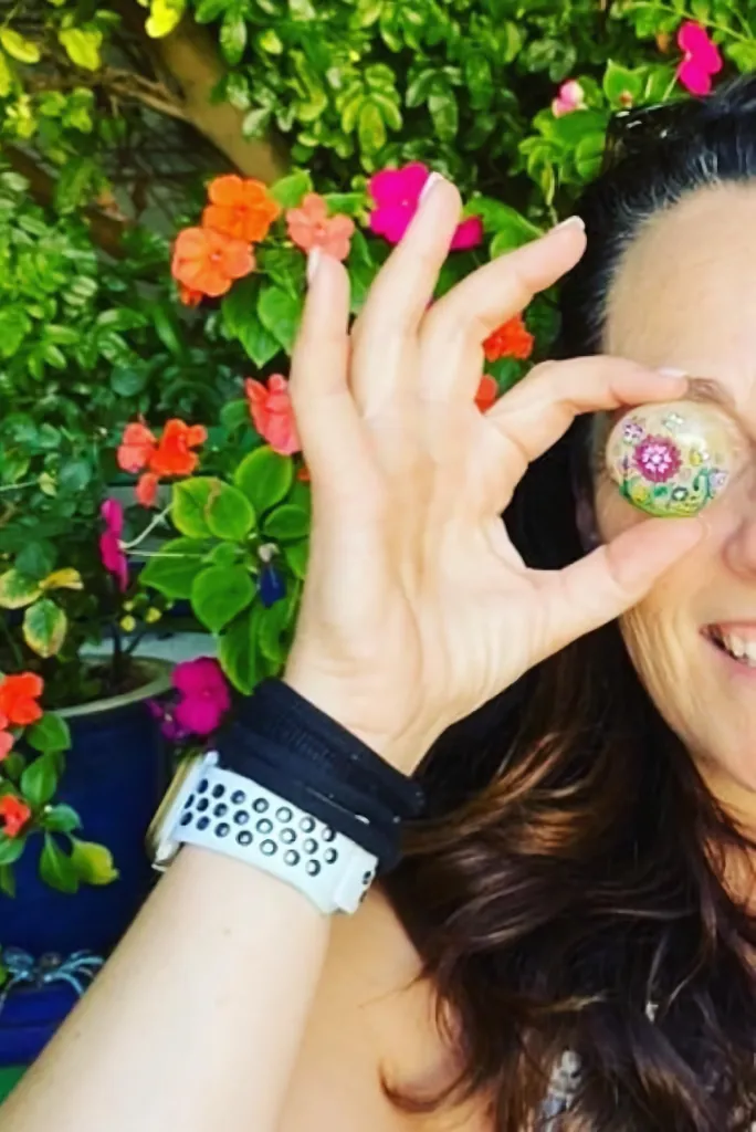 Image of Sam, owner of Pocket Pebbles, holding flower painted pebble in front of one, face showing only half the face, backdrop of colourful flowers and green leaves