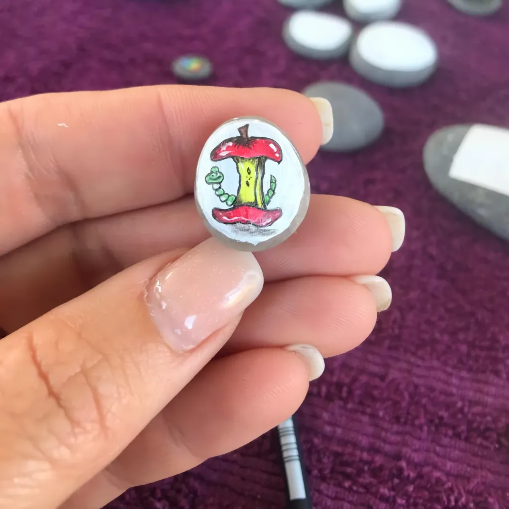tiny apple core and worm, colourful painting on a tiny pebble balanced on the finger tips of a lady's hand