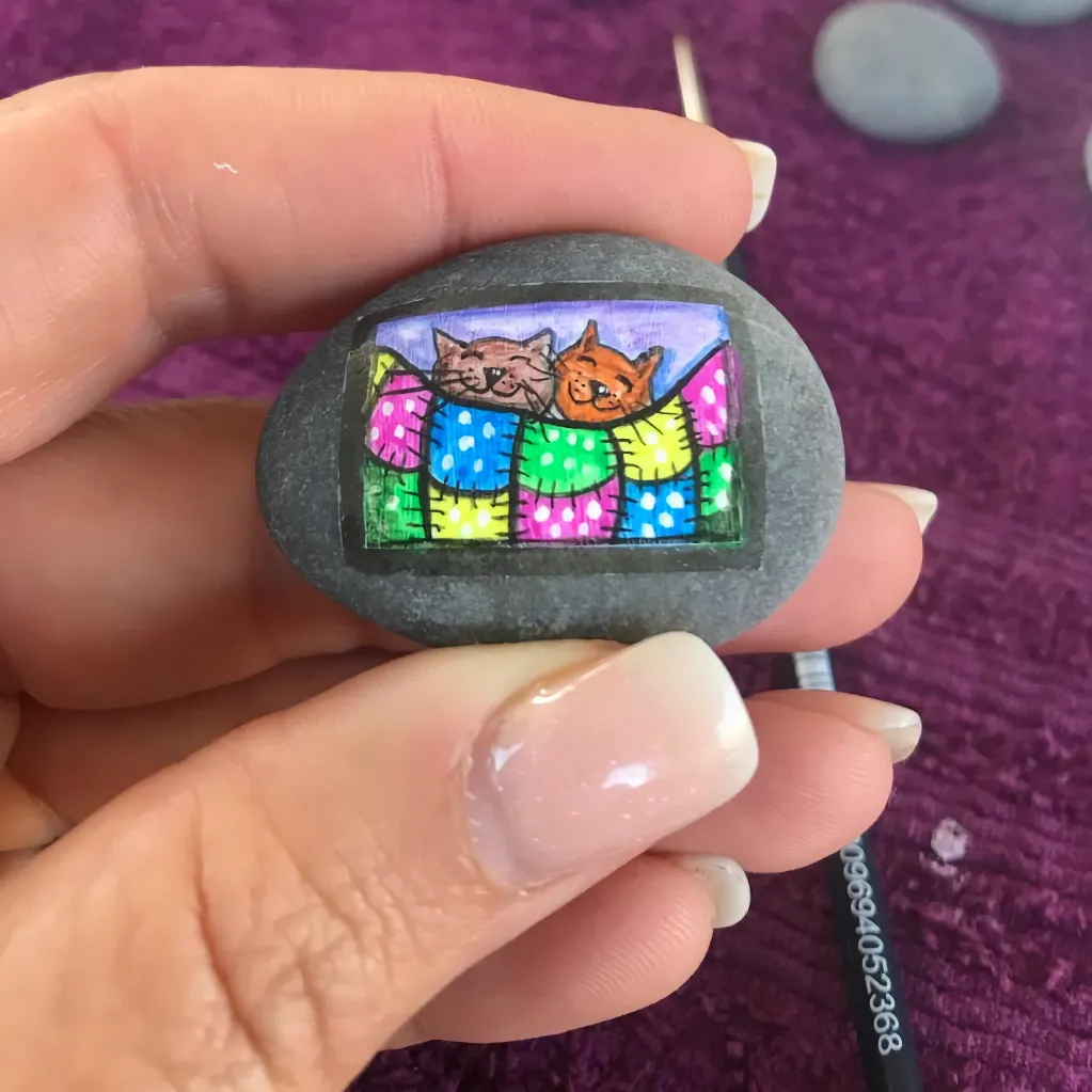 two cats under a colourful patchwork blanket painted on a grey stone held between thumb and forefinger of lady's hand