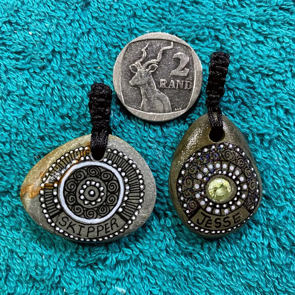 two painted pebble dog tags next to a two Rand coin to illustrate size on teal towel