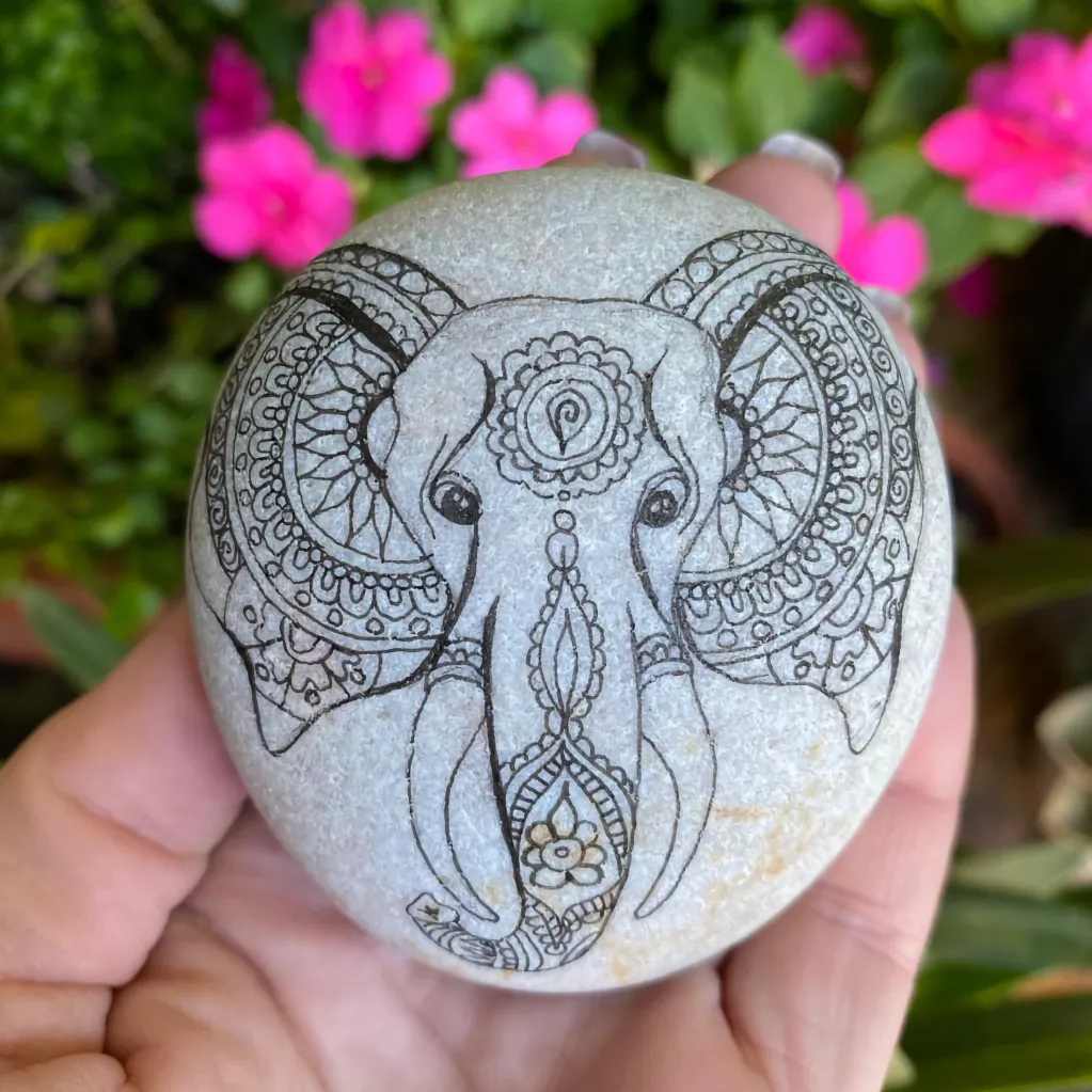 Asian type painted elephant face painted on a pale stone and held by a lady's hand in front of a pink flowered green bush
