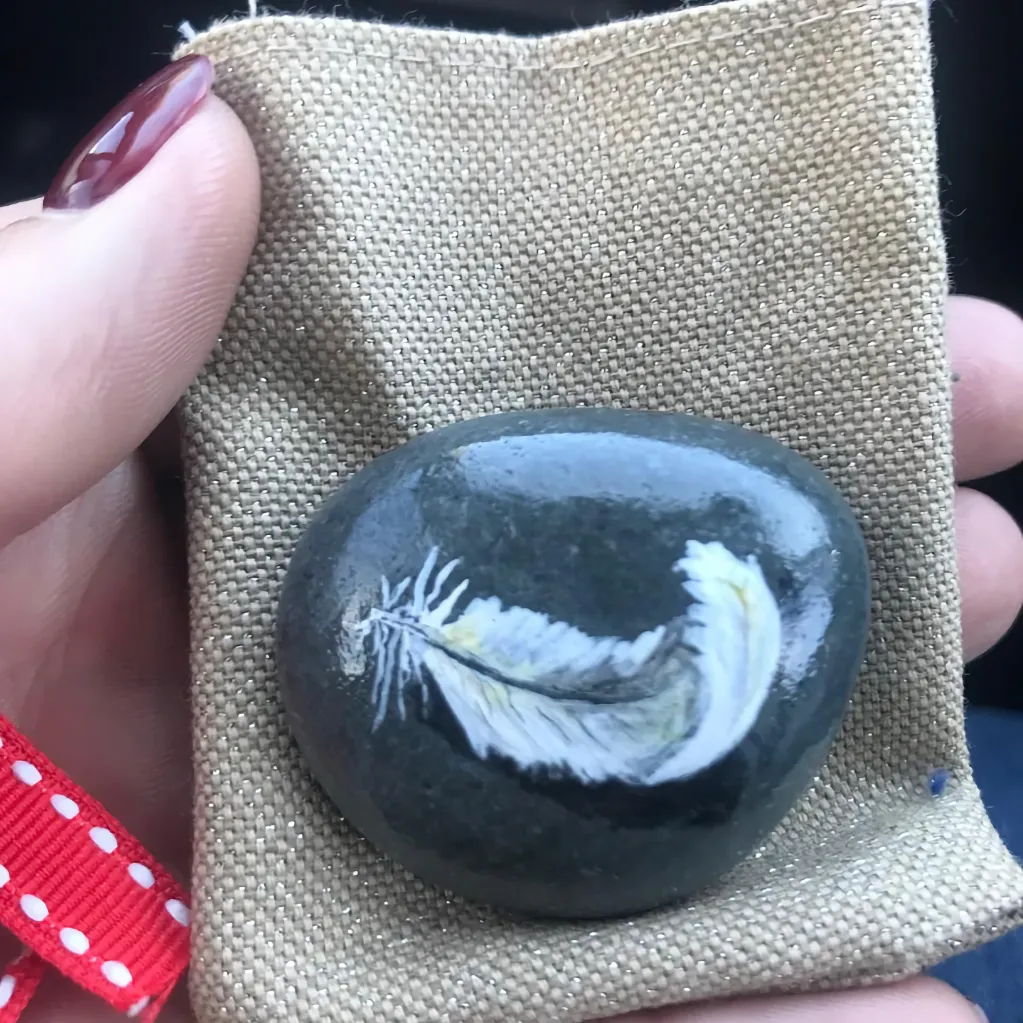 peaceful feather painted on dark stone in front of hand holding pebble atop a sack like gift bag